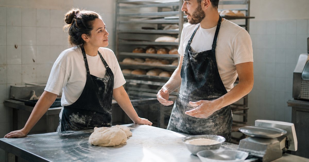 Two coworkers in aprons standing and talking beside a stainless steel counter in a bakery back room at midday, trays of bread on a rolling rack behind them, flour dust in the air, soft natural light from a high window