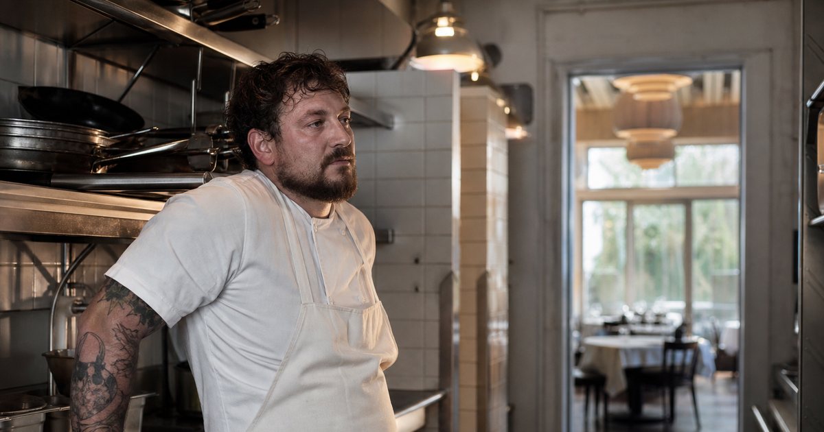 A tired line cook in a white apron leaning against a stainless steel prep counter in a quiet restaurant kitchen just before opening, clean pans stacked on a shelf behind him, warm overhead lamps and pale daylight coming through a doorway into an empty dining room
